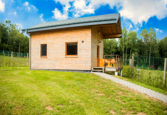 Cozy wooden chalet named Rogery 64 in Belgium, set in green fields with forest and blue sky all around.