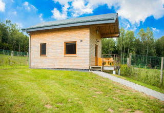 Knus houten chalet Rogery 64 in België, omringd door groene velden en bos, onder een stralend blauwe lucht.