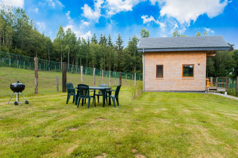 Chalet Rogery 64 in België met houten huisje, tuinset, barbecue en grasveld naast een bosrijke omgeving.