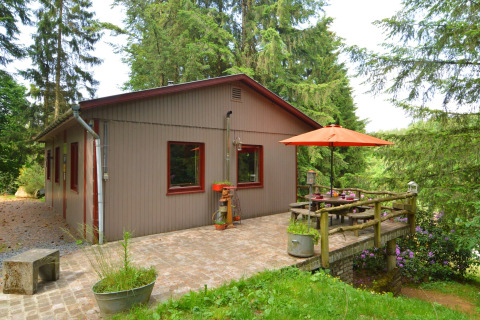 Udsigt til chalet 'Flons et Roulotte' i Belgien, med terrasse, bord, parasol og omgivet af grønne træer.