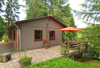 Udsigt til chalet 'Flons et Roulotte' i Belgien, med terrasse, bord, parasol og omgivet af grønne træer.
