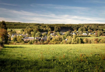 View from chalet Rogery 64 in Belgium, overlooking green fields, forest, and a quaint village beyond.