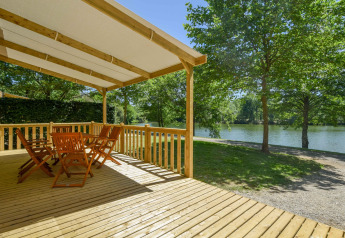 Überdachte Holzterrasse mit Sitzgruppe und Blick auf See und Bäume am Mobilheim Cottage „6p“ in Frankreich.