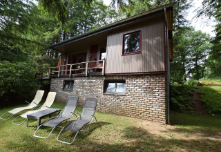 Chalet Flons et Roulotte i Belgien omgivet af skov, med fire liggestole på græsset foran huset.