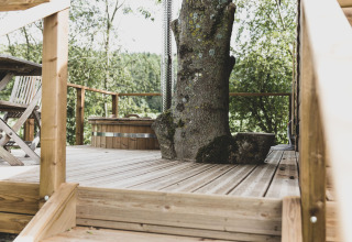 Terrazza della casa sull’albero Bird Nest in Belgio con un grande albero e mobili in legno nel verde.