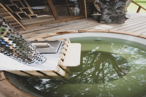 Outdoor wooden hot tub with greenish water on deck of 'Bird Nest' tree house in Belgium, tree reflections.