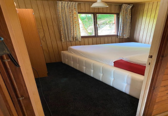 Slaapkamer in houten chalet met groot wit bedframe, ramen en bloemige gordijnen in België.