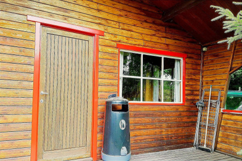 Uitzicht op chalet La Bressine in België met houten deur, rood kozijn, raam, vuilnisbak en veldbed.