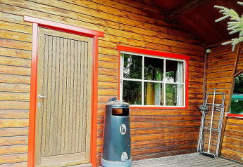 Zicht op chalet La Bressine in België met houten deur, rood-kaderraam, vuilbak en veldbed.