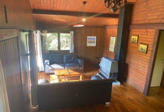 Woonkamer in chalet La Bressine in België met houten wanden, banken, kachel en grote ramen.