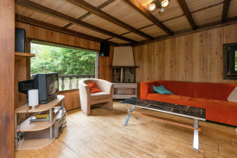 Houten woonkamer in lodge Le Chession in België met oranje bank, tv en uitzicht op het bos.
