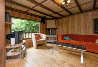 Houten woonkamer in lodge Le Chession in België met oranje bank, tv en uitzicht op het bos.