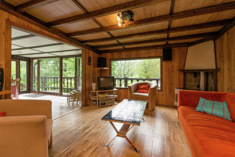 Woonkamer in lodge 'Le Chession' in België met houtafwerking, veel ramen en zicht op het bos.