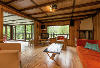 Woonkamer in lodge 'Le Chession' in België met houtafwerking, veel ramen en zicht op het bos.
