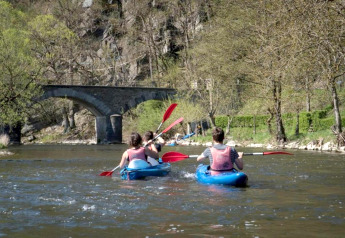 Personen paddeln in Kajaks auf einem Fluss bei Le Chession in Belgien mit einer Steinbrücke im Hintergrund.