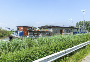 Casa galleggiante con turbina eolica e vasi di piante sul tetto, vista da una strada in Belgio.