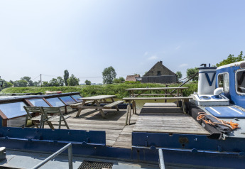 Ponte esterno della houseboat 'St Antoine Isera' in Belgio con panche, tavoli in legno e vista rurale.
