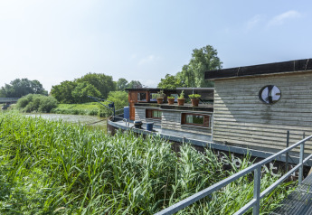 Casa galleggiante 'St Antoine Isera' ormeggiata lungo il fiume tra vegetazione rigogliosa, Belgio.
