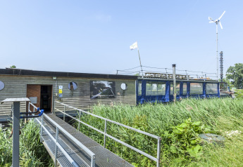 Houseboat St Antoine Isera con turbina eolica, vegetazione e rampa in una giornata di sole in Olanda.