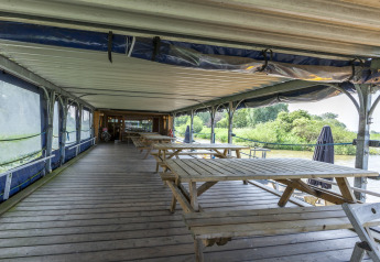 Terrazza coperta in legno con tavoli da picnic sulla houseboat St Antoine Isera in Belgio, vista sul verde.