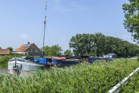 Husbåd ved navn St Antoine Isera fortøjet ved en grøn kanal i Belgien med hus og træer i baggrunden.