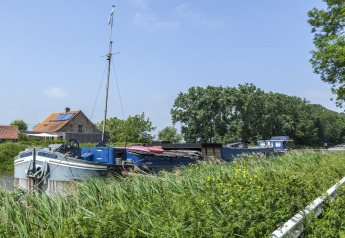 Houseboat chiamato St Antoine Isera attraccato su un canale verde in Belgio, con casa e alberi dietro.