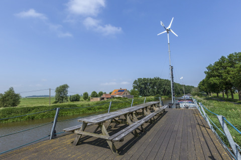 Træborde og bænke på husbåden St Antoine Isera i Belgien med vindmølle og landskab i baggrunden.