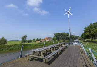 Træborde og bænke på husbåden St Antoine Isera i Belgien med vindmølle og landskab i baggrunden.