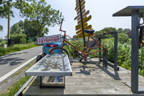Cykel og skilte på en husbåd ved vejen i Belgien, med grønt landskab og skilte med retninger.