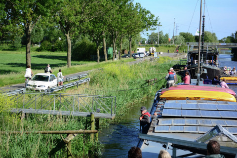 Hausboot 'St Antoine' in Belgien, mit Menschen an Bord und Radfahrern sowie Autos auf der Flussstraße.