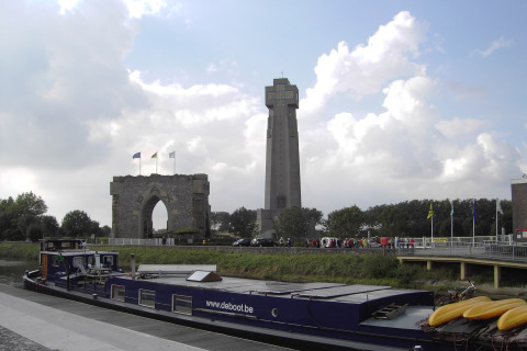 Das Hausboot „St Antoine“ liegt in Belgien an einem Kanal, im Hintergrund ein Denkmal und hoher Turm.