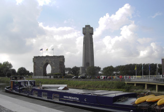 Das Hausboot „St Antoine“ liegt in Belgien an einem Kanal, im Hintergrund ein Denkmal und hoher Turm.