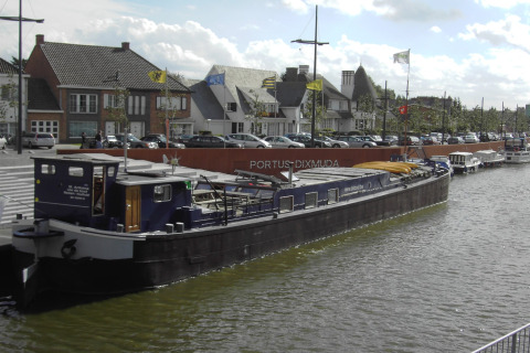Hausboot St Antoine liegt am Kanal in Belgien, mit Stadtgebäuden und geparkten Autos im Hintergrund.