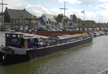 Huspram ved kajen i Belgien, omgivet af moderne bygninger og parkerede biler, med PORTUS DIXMUDA baggrunden.