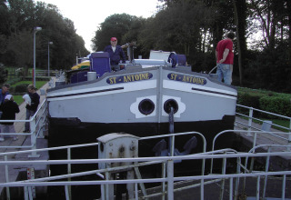 Personen auf und um das Hausboot 'St Antoine' in einer Schleuse in Belgien, umgeben von Bäumen.