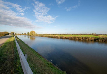 Udsigt fra husbåden 'St Antoine' i Belgien, der viser en rolig kanal, grønne marker og en blå himmel.