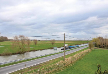 Udsigt fra husbåden 'St Antoine' i Belgien, med grønne marker, veje og en flod under en overskyet himmel.
