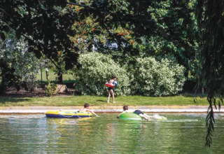 Kinder spielen mit Schwimmringen im Wasser vor dem Hausboot St Antoine in Belgien unter Bäumen.