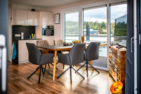 Interno di una moderna houseboat con cucina, zona pranzo e grandi finestre affacciate sul lago Bärwalder See.