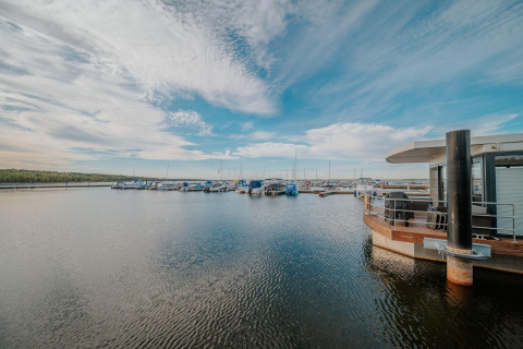 Vista de Houseboat am Bärwalder See amarrado en un muelle junto a barcos y cielo con nubes dispersas.