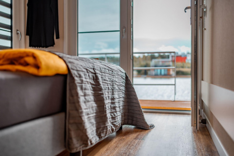 Bedroom with bed and blanket, sunlight pouring in from an open door with lake view on Houseboat am Bärwalder See.