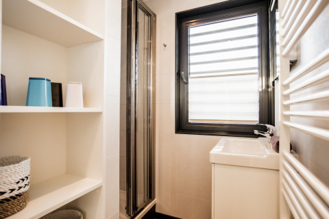 Lyst og moderne badeværelse på en husbåd med brusekabine, vindue, åbne hylder og håndvask.