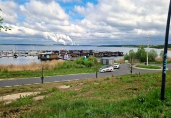 Blick auf Houseboat am Bärwalder See mit Hausbooten, Autos, Straße und rauchendem Werk im Hintergrund.