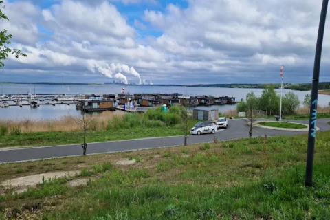 Udsigt over Houseboat am Bärwalder See med husbåde, biler, vej og skytende fabrik i baggrunden.