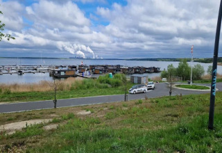 View of Houseboat am Bärwalder See showing houseboats, cars, a road, and smoking factory in the distance.