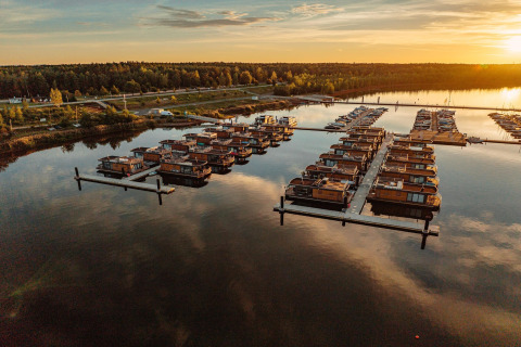 Solnedgangsbillede af Houseboat am Bärwalder See, flere husbåde på vandet nær en skovklædt kyst.