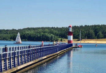 Blick auf einen Steg mit blauem Geländer, Leuchtturm und Segelbooten bei Houseboat am Bärwalder See an einem sonnigen Tag.