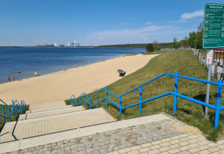 Blue-railed steps lead to a sandy beach by Houseboat am Bärwalder See with swimmers and clear weather.