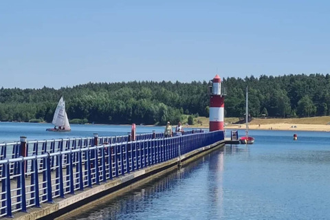 Casa flotante en Bärwalder See con muelle azul, faro rojo-blanco, velero y orilla arenosa soleada.