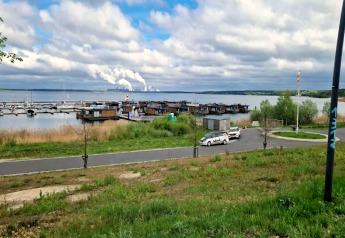 Hausboot am Bärwalder See mit Blick auf Wasser, Hafenanlagen, Autos und Schornsteine im Hintergrund.